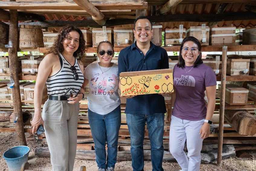 Four people standing in an outdoor setting with wooden structures and beehive boxes in the background. 4