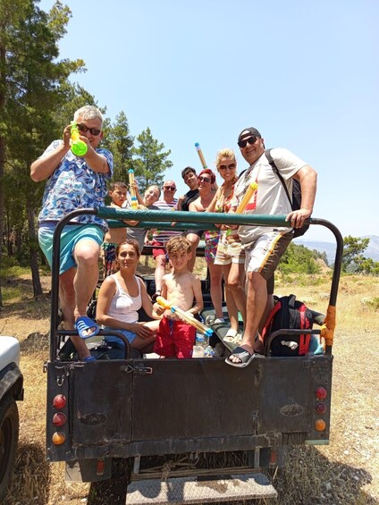 A group of people in a vehicle with a trailer, some holding water bottles, in a natural outdoor setting. 5