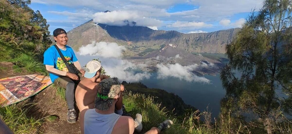 Two people sitting on a mountain ledge overlooking a valley with a lake and clouds above. 1