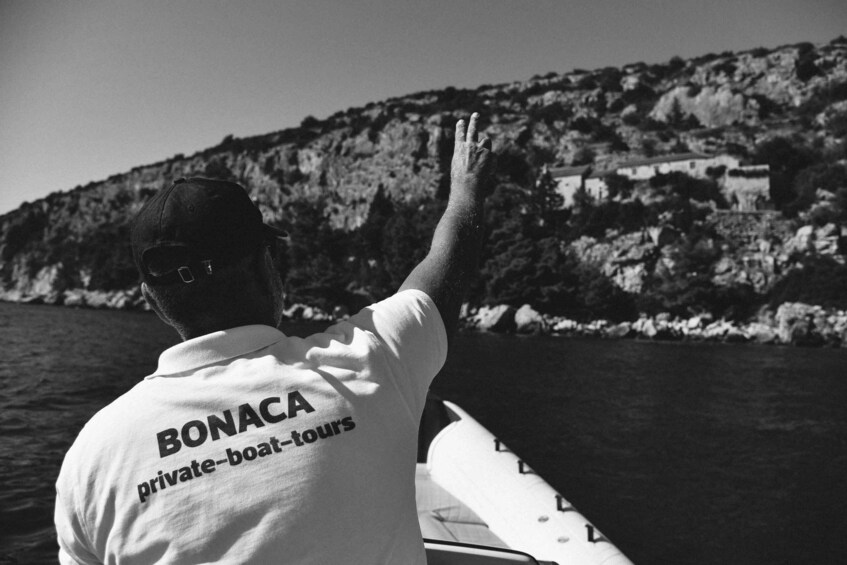 A person on a boat wearing a shirt with "BONACA private-boat-tours" text, with a rocky coastline in the background. 4