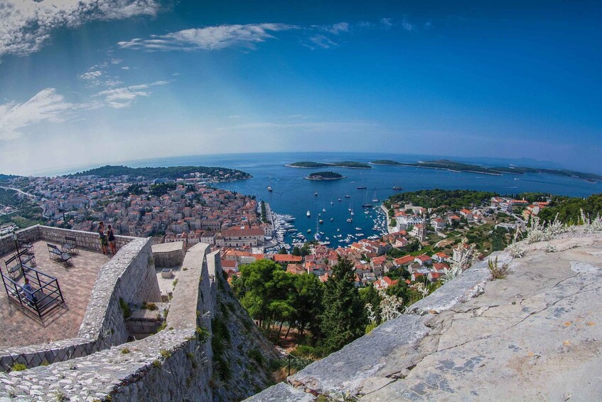 A coastal town with a historic fortification, a marina with boats, and a clear blue sky. 1