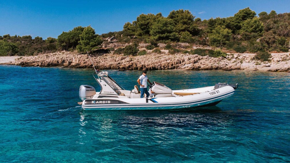 A white motorboat with 'KARDIS' and '1550 ST' on it, on clear blue water with a rocky shoreline and trees in the background. 2