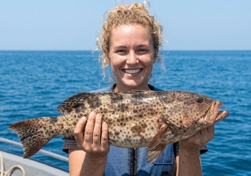 A person holding a fish with a spotted pattern, standing on a boat with a blue ocean in the background. 1