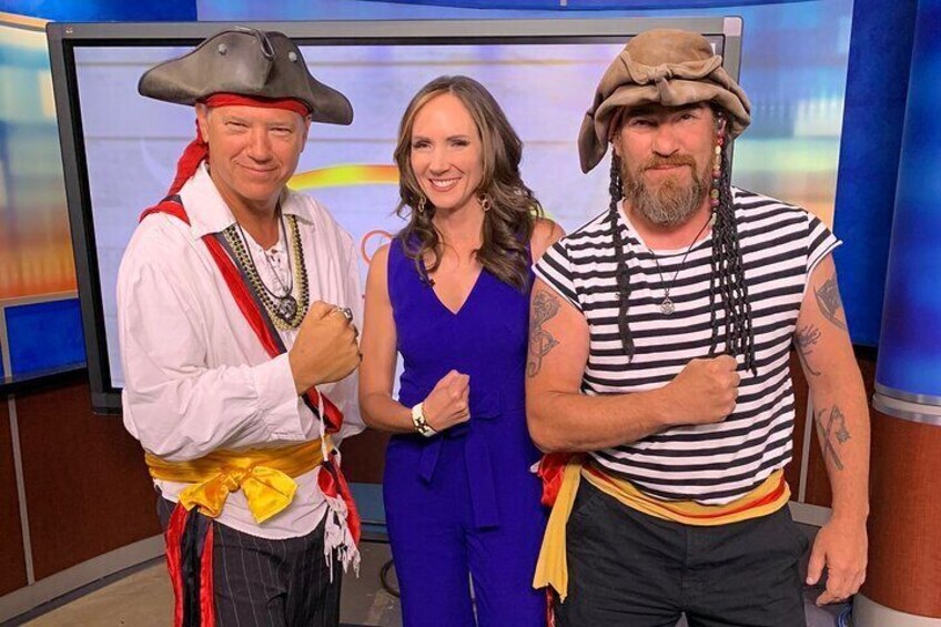 Three people in costumes, a man in a pirate outfit and a woman in a blue dress, standing in a studio with a blue background. 4