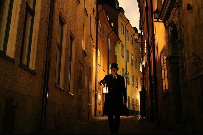 A person walking down a narrow street flanked by buildings with large windows and street lamps. 5