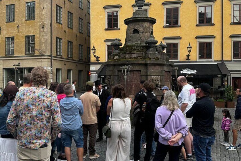A group of people gathered around a historic fountain in a public square with yellow buildings in the background. 2