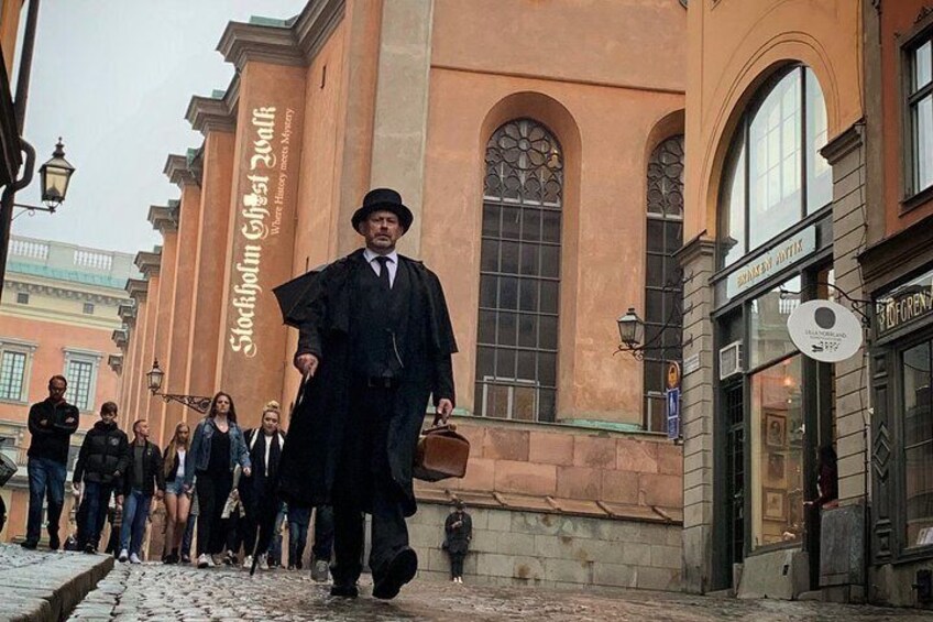 A man in a black suit and hat walking on a cobblestone street with a crowd behind him. 1