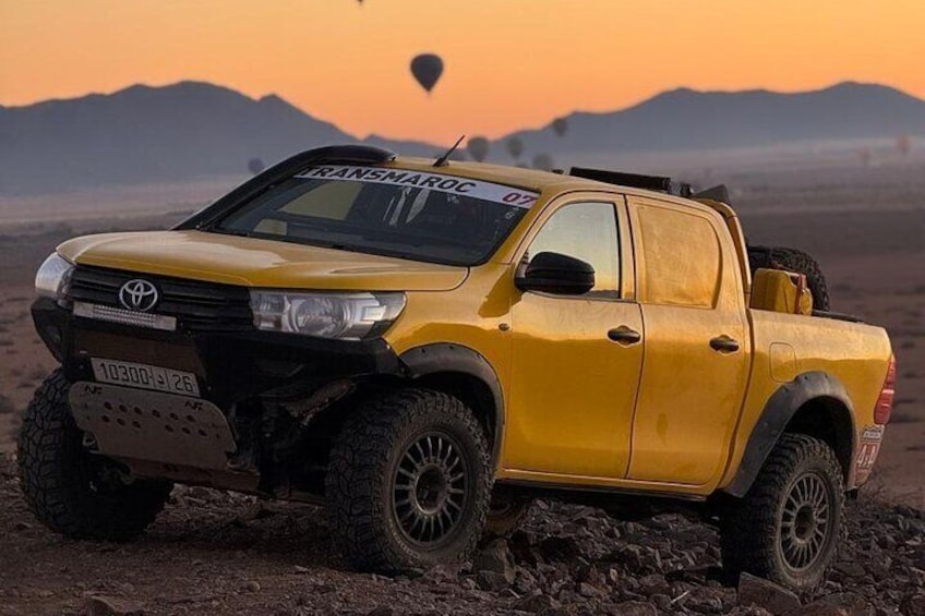 A yellow Toyota pickup truck with off-road tires and a roof rack, parked on a rocky terrain with a sunset in the background. 1