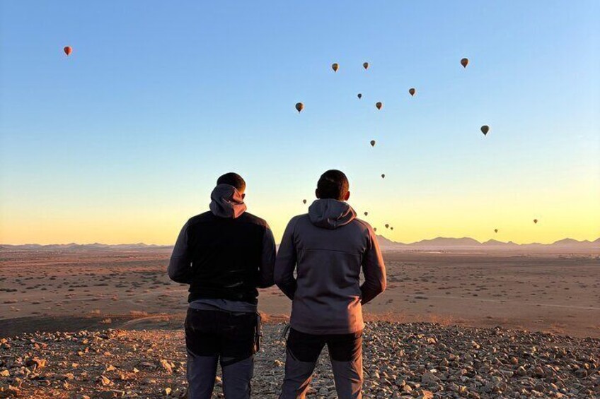 Two people standing on a rocky terrain watching hot air balloons in the distance. 2