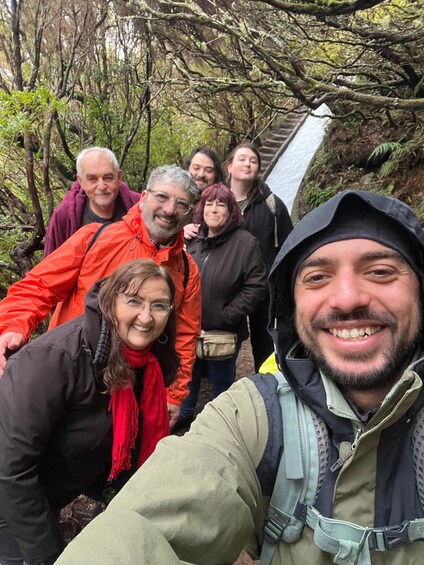 A group of people taking a selfie in a forested area with a wooden bridge in the background. 1