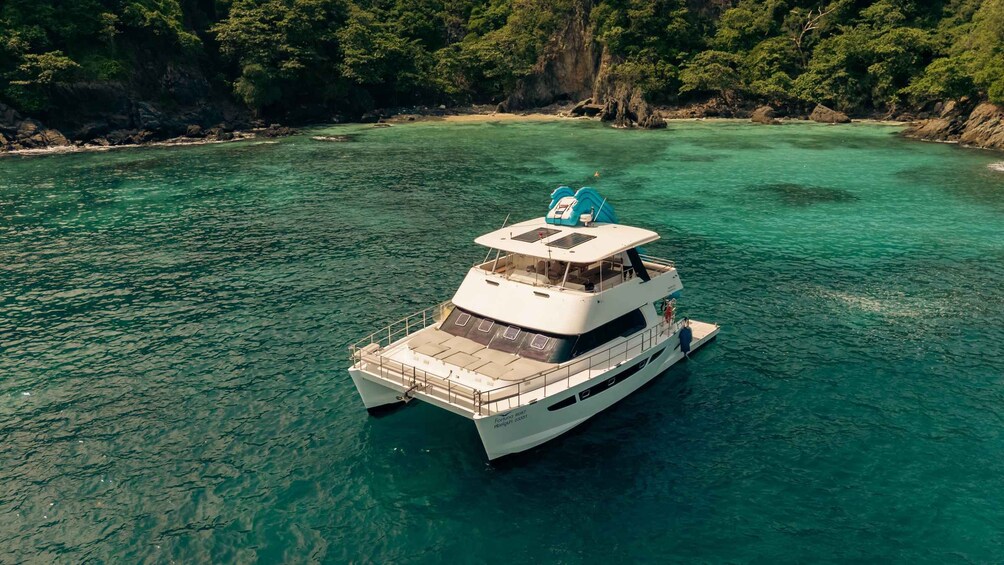 A white boat with a cabin and a blue canopy on top, floating on clear turquoise water near a rocky coastline with dense green foliage. 1