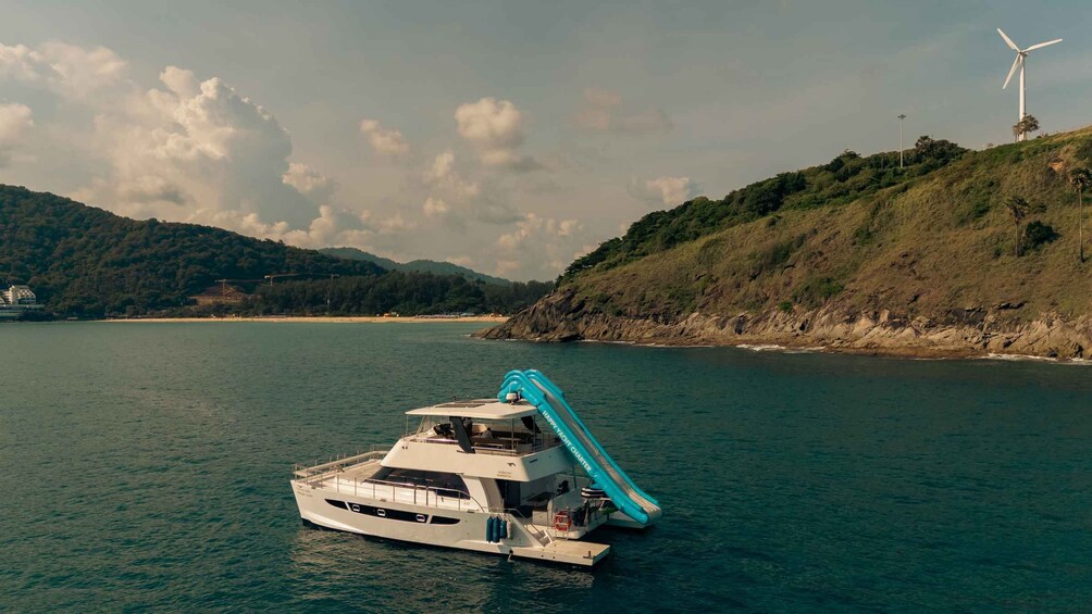 A yacht with a water slide on a calm sea, a wind turbine in the background, and a hilly coastline. 5
