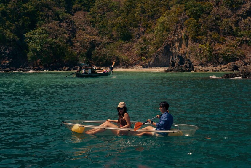 Two people paddling a transparent boat on clear turquoise water, with a rocky coastline and a boat in the background. 2