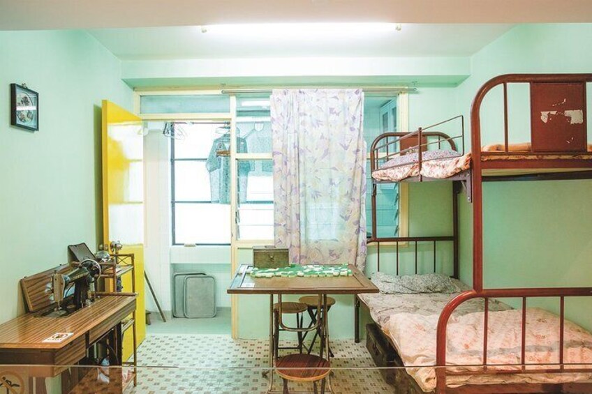 A dormitory room with bunk beds, a desk with a typewriter, a window with a curtain, and a radiator. 5