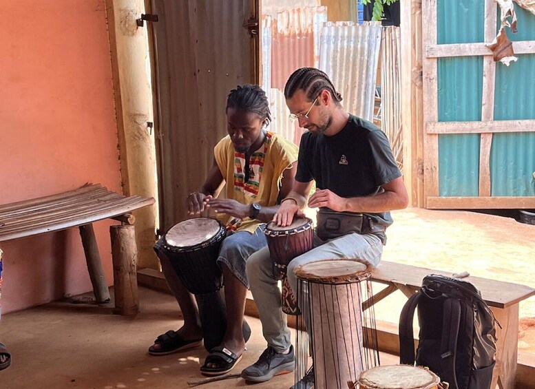 Two individuals playing drums in an outdoor setting with a wooden bench and corrugated metal walls. 2
