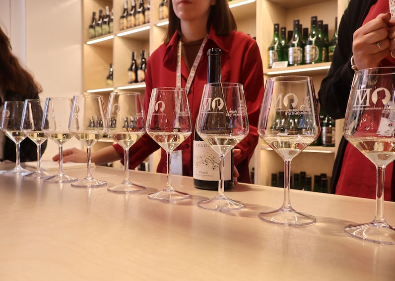 Une dégustation de vins, avec de nombreux verres à vin disposés sur une table, une personne tenant une bouteille de vin, et des étagères remplies de bouteilles de vin en arrière-plan. 3