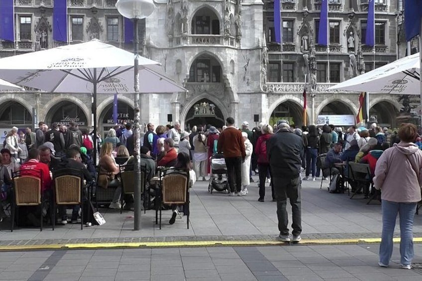 A crowded outdoor café with people sitting at tables under umbrellas, a historic building in the background, and a yellow line marking the edge of the pavement. 2