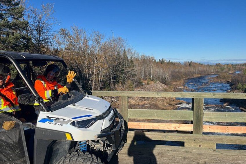 A white off-road vehicle with 'XP' and 'KINETTE' on the front, parked on a wooden bridge with a river and trees in the background. 4