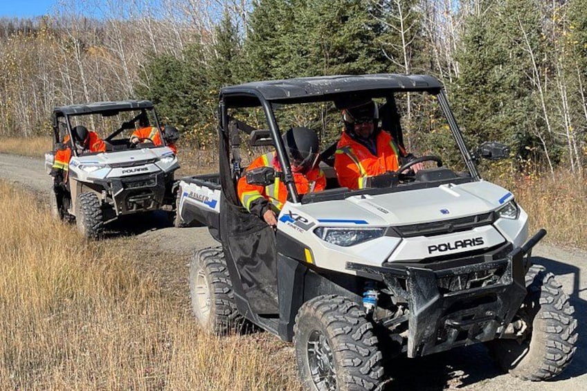 Two Polaris all-terrain vehicles on a dirt road with forest in the background. 1