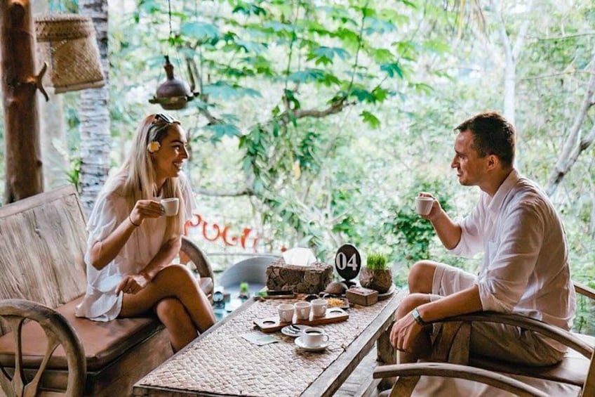 Two people sitting at a wooden table outdoors, holding cups, with a rustic setting including a wicker chair and a stone wall in the background. 5