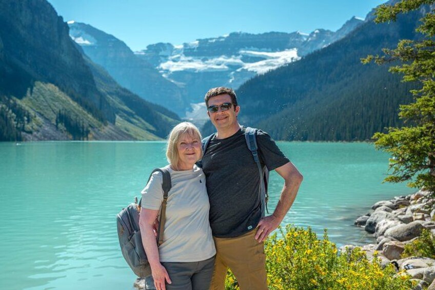 Twee personen staan bij een turquoise meer, met bergen op de achtergrond. 3