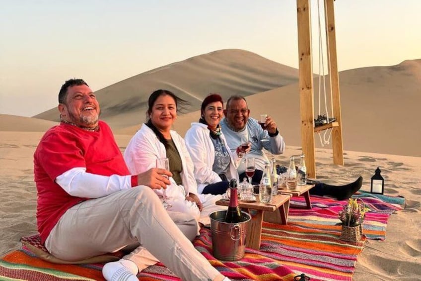 A group of people sitting on a blanket in a desert with sand dunes in the background. 1