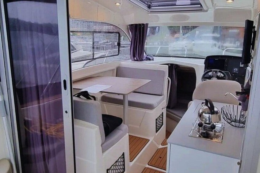 A compact boat interior with a dining area, a control panel, and a window overlooking the water. 4