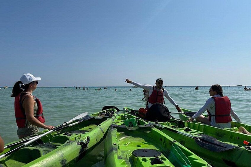 A group of people in green kayaks on a calm body of water, wearing life jackets. 2