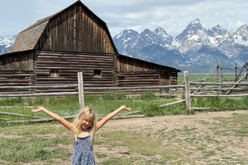 A child in a blue dress with arms outstretched in front of a rustic wooden barn with snow-capped mountains in the background. 1