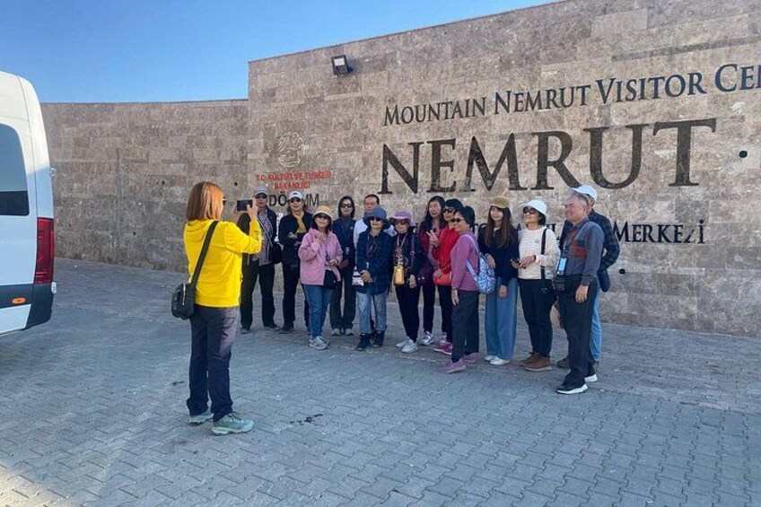 A group of people standing in front of a wall with the text 'MOUNTAIN NEMRUT VISITOR CENTER' and 'NEMRUT' on it. 1
