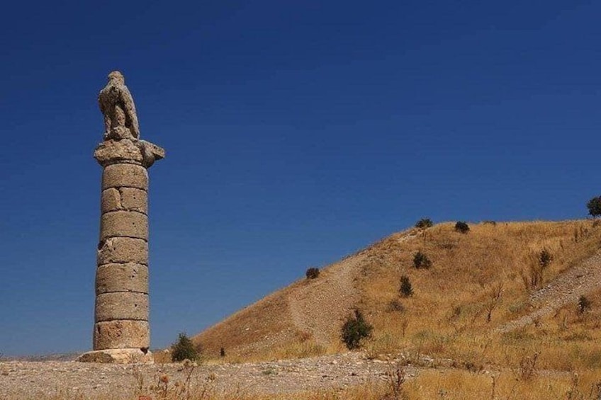 A stone column with a statue on top, set against a clear blue sky and a dry, grassy hill. 3