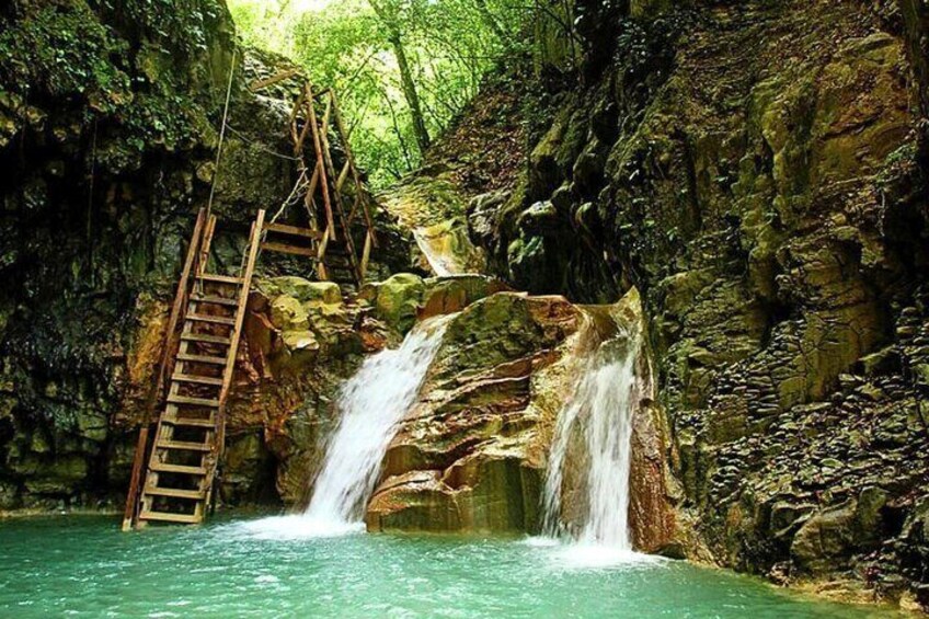 A waterfall with a wooden ladder leading up to it, surrounded by rocky terrain and greenery. 1