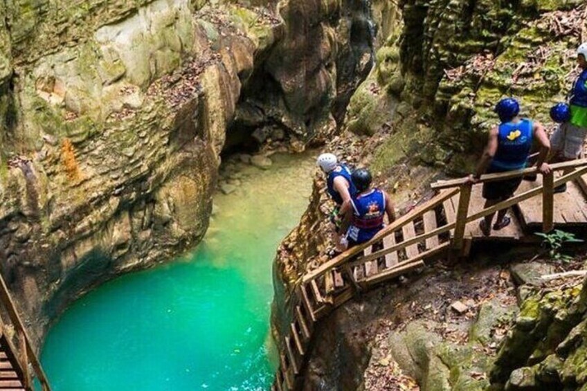 A group of people in blue helmets and life vests on a wooden bridge over a turquoise pool surrounded by rocky terrain. 3