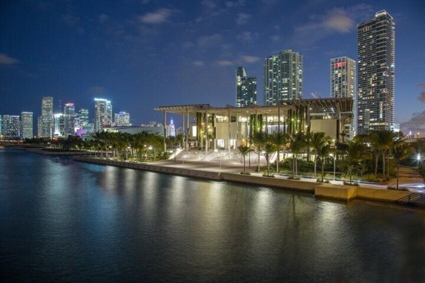 A waterfront cityscape with modern buildings, a well-lit public space, and a calm body of water reflecting the lights. 5