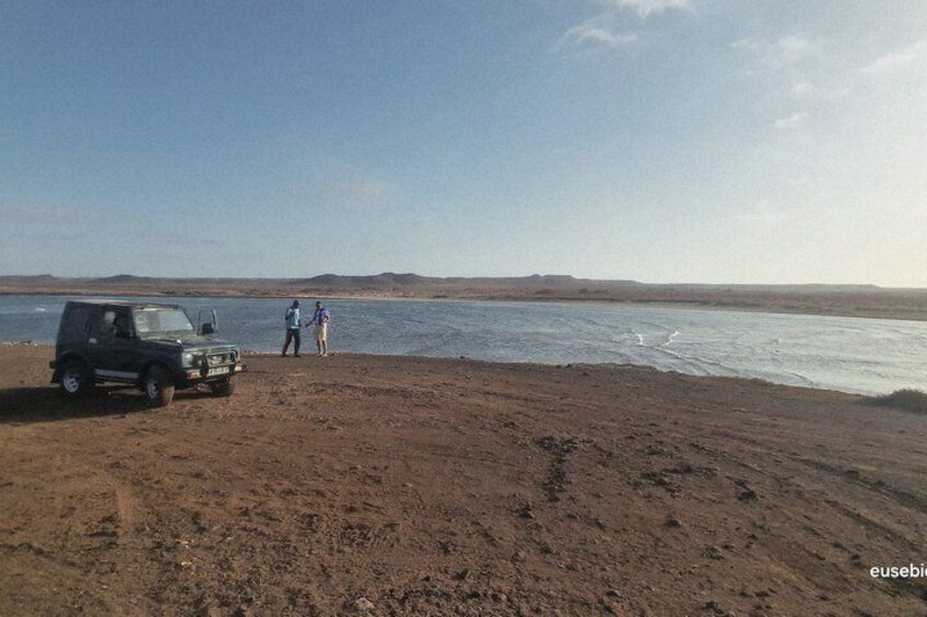 A desert landscape with a body of water, a parked vehicle, and two people standing by the water's edge. 2