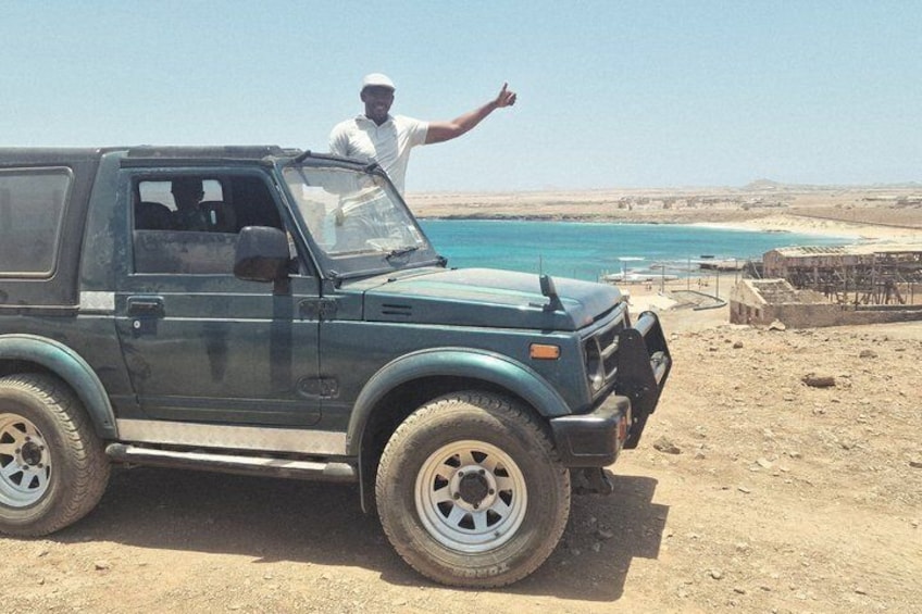 A green off-road vehicle parked on a sandy beach with a person standing in the driver's seat. 1