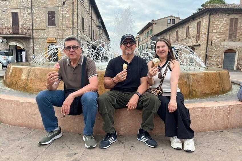 Three people sitting on a bench near a fountain in a historic town square. 1