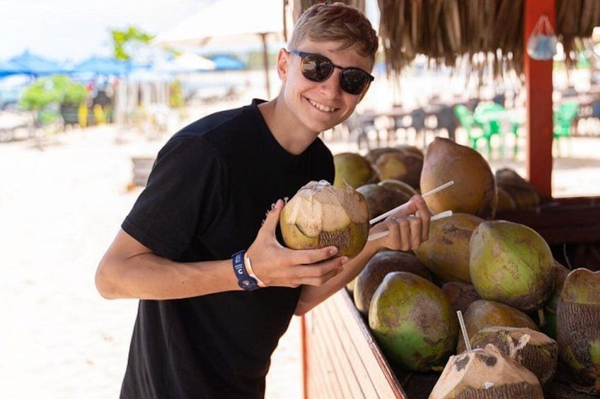A person holding a coconut with a straw, standing next to a pile of coconuts. 3