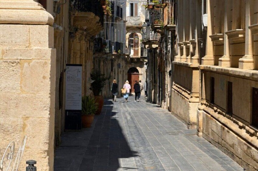 A narrow street with stone buildings, potted plants, and a few pedestrians. 3