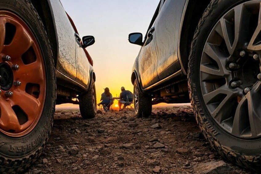 A vehicle's wheels on a dirt path with two people sitting at a campfire. 1