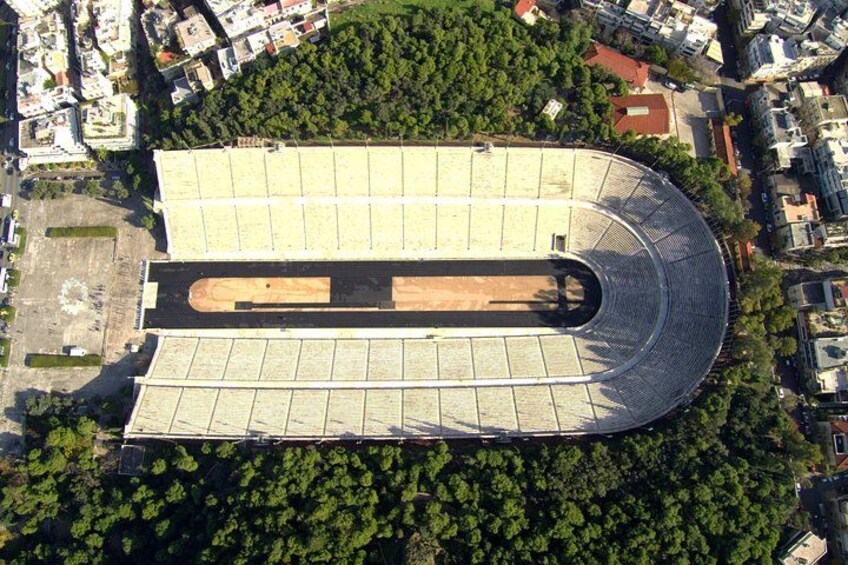 An aerial view of a stadium with a curved roof, surrounded by dense greenery and urban buildings. 3