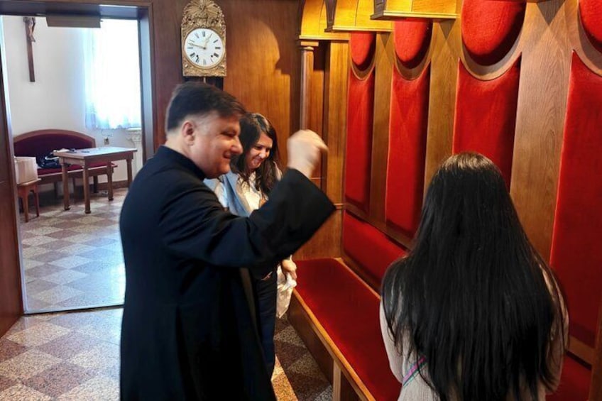 A person in a black coat standing in a room with red upholstered seating, a wooden wall, and a clock. 1