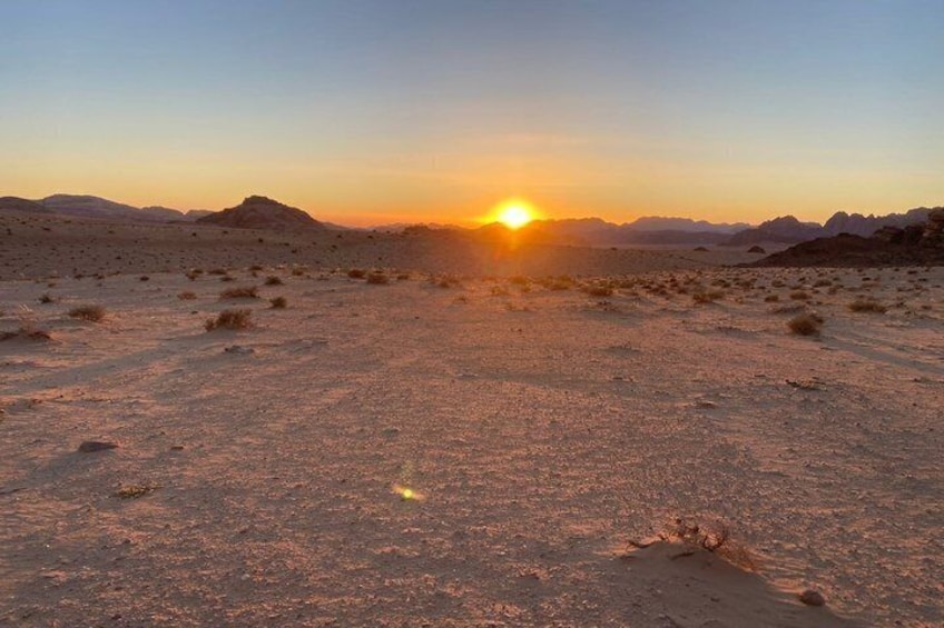 A desert landscape at sunset with a visible sun, rocky terrain, and sparse vegetation. 4