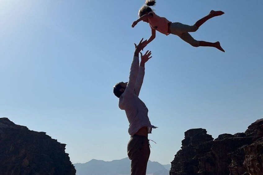 A person jumping off a cliff with another person mid-air, rocky cliffs, and mountains in the background. 3