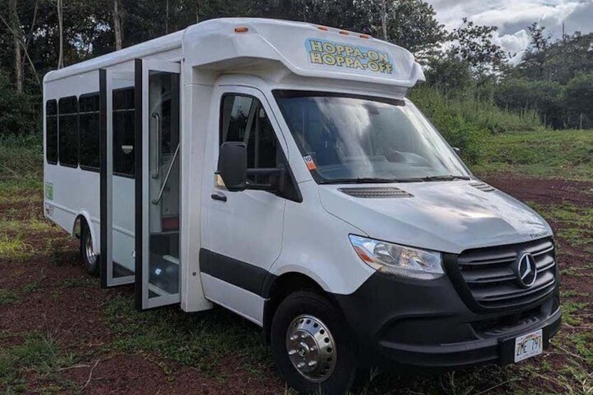 A white Mercedes van with a 'HOPPA-ON HOPPA-OFF' sign, parked on a dirt road with greenery in the background. 4