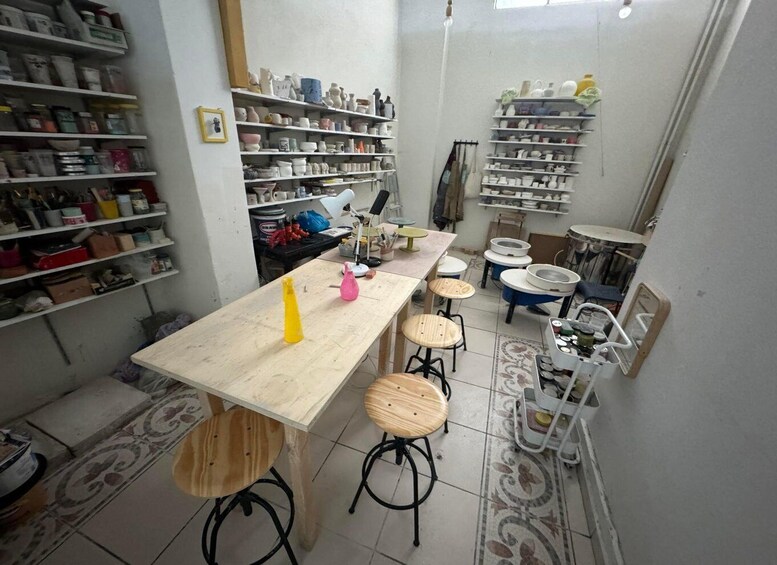 A pottery studio with shelves of ceramics, a workbench, stools, and various pottery tools. 1