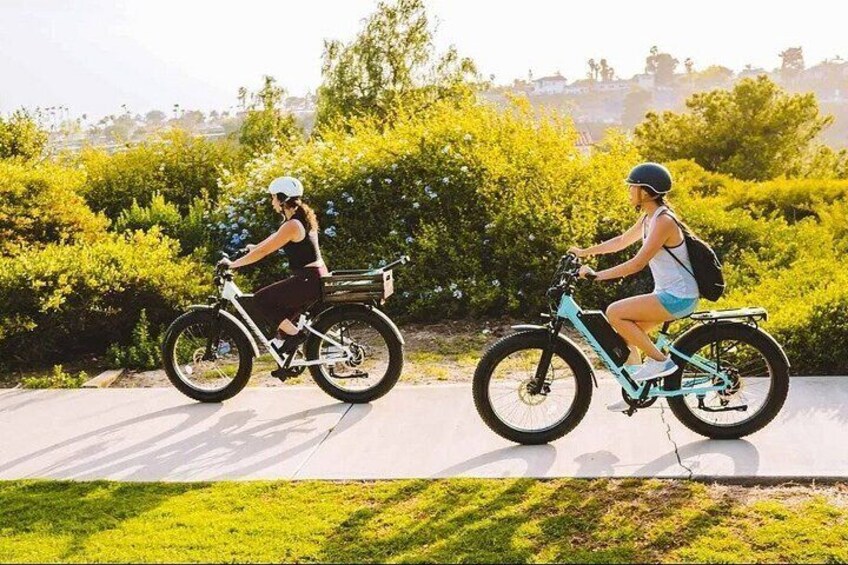 Two people riding electric bicycles on a paved path with greenery and trees in the background. 3