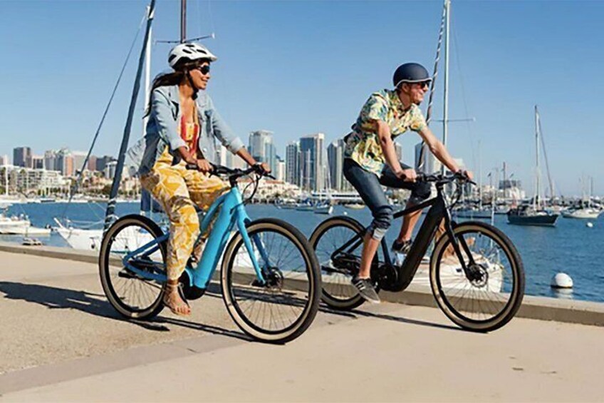 Two people riding bicycles by a waterfront with boats and a city skyline in the background. 5