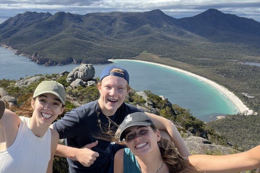 A group of people taking a selfie with a scenic view of a coastline and mountains in the background. 1