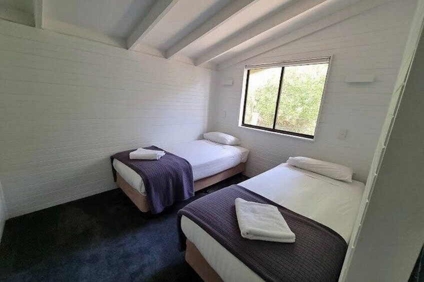 A small, compact room with two beds, a window, and white walls. 3